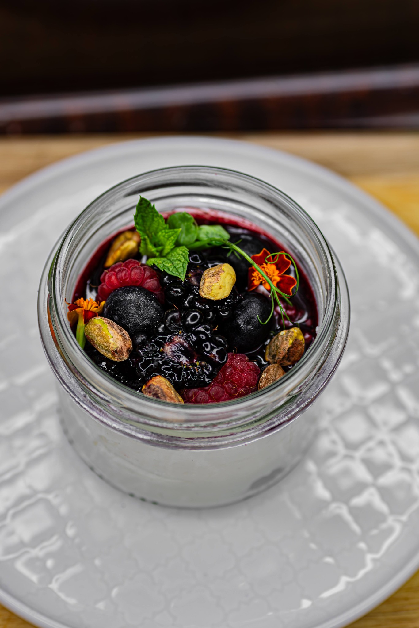 Deser w słoiku z jogurtem, sosem jagodowym, malinami, jeżynami, borówkami, pistacjami i miętą na białym talerzu.