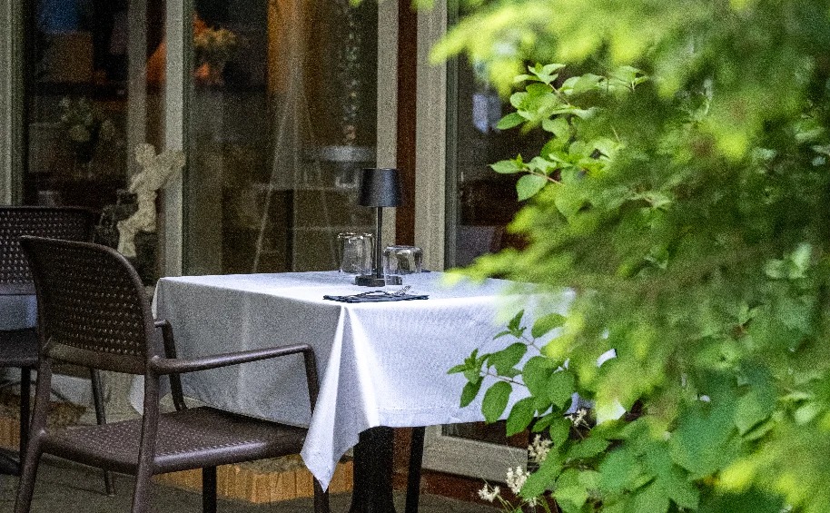 Stół nakryty białym obrusem na zewnątrz restauracji, z widocznymi szklankami, sztućcami i lampką, częściowo zasłonięty zielenią.