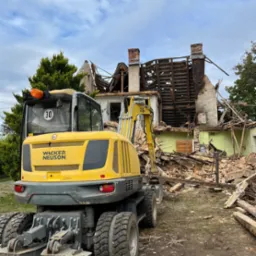 Żółta koparka Wacker Neuson na tle zrujnowanego budynku z widocznymi elementami konstrukcji, cegłami i kominem, w tle drzewa i pochmurne niebo.