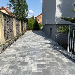 SGE - Nowo ułożona, szara kostka brukowa na podjeździe, z rzędem betonowych donic z pomarańczowymi kwiatami wzdłuż ażurowego ogrodzenia.