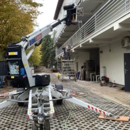 SGE GRZEGORZ GIL - Podnośnik koszowy DINO SYSTEM ustawiony na ażurowej nawierzchni, pracownicy na rusztowaniu i balkonie budynku w tle.