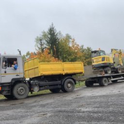 SGE GRZEGORZ GIL - Ciężarówka MAN z żółtą skrzynią ładunkową ciągnąca lawetę z żółtą koparką kołową na tle jesiennego krajobrazu.