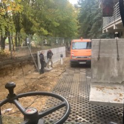 SGE GRZEGORZ GIL - Widok z kabiny maszyny na plac budowy z wykopem, pracownikami i pomarańczowym samochodem dostawczym na tle drzew i budynku, widoczne betonowe elementy konstrukcyjne.