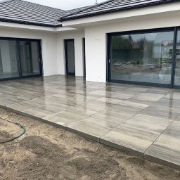 SGE - Świeżo ułożony, mokry taras z szarych, prostokątnych płyt imitujących kamień, przylegający do białego domu z dużymi przesuwnymi drzwiami tarasowymi i czarnym dachem, widoczny wąż ogrodowy na skraju.