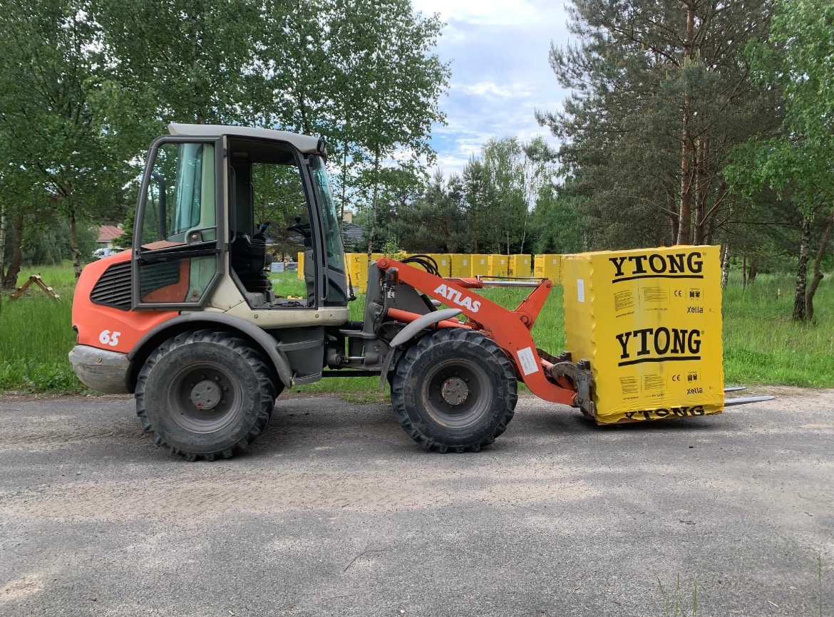 Pomarańczowy ładowacz teleskopowy Atlas przewozi paletę żółtych bloczków Ytong na terenie budowy.
