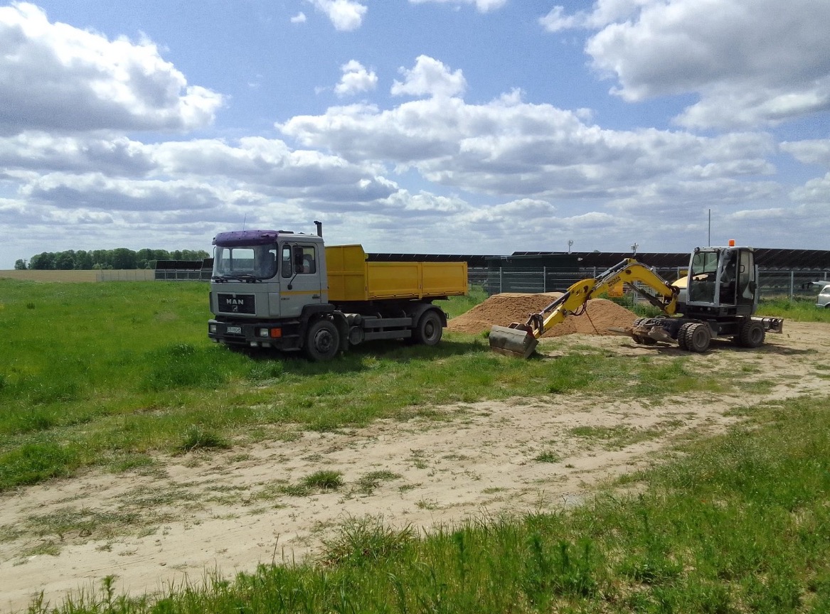 Żółto-szara wywrotka MAN i koparka kołowa przy pryzmie piasku na trawiastym terenie, w tle farma fotowoltaiczna i błękitne niebo z chmurami.