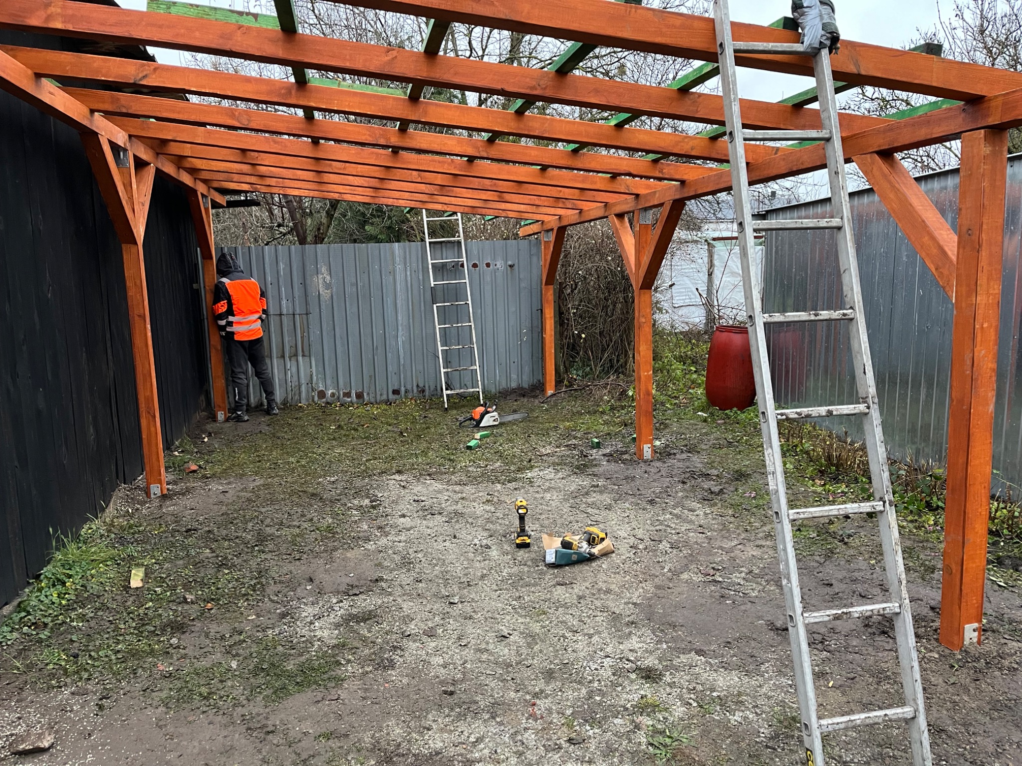 Drewniana konstrukcja z belek w kolorze brązowym, częściowo zadaszona, w trakcie montażu na zewnątrz, widoczny pracownik w kamizelce odblaskowej oraz narzędzia i drabiny.