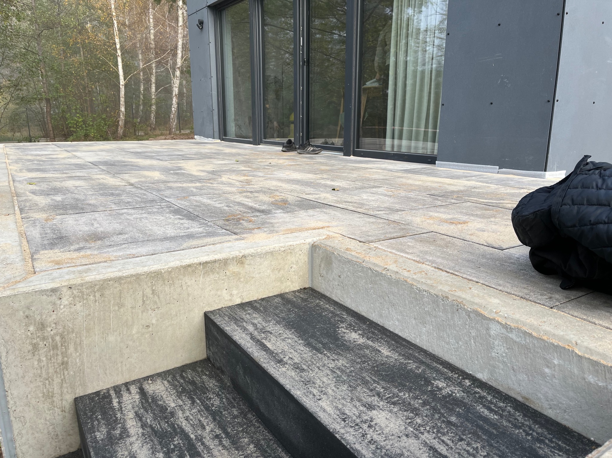Nowoczesny taras z szarej kostki brukowej z betonowymi schodami, widoczne buty i torba przy przeszklonych drzwiach domu, tło stanowią brzozy.