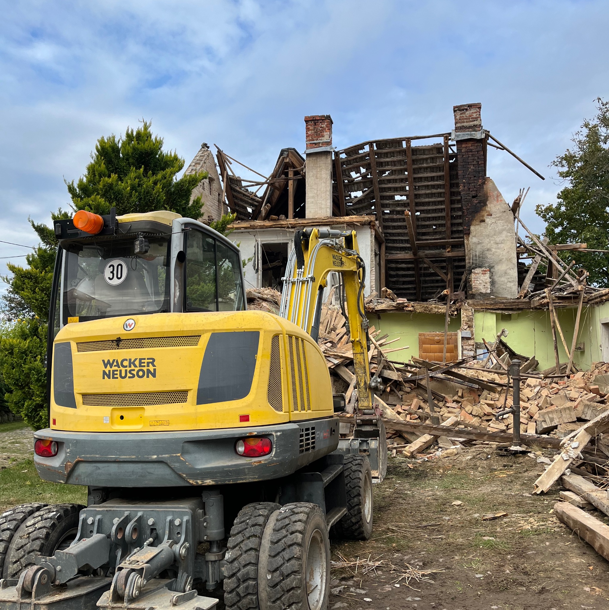Żółta koparka Wacker Neuson na tle zrujnowanego budynku z widocznymi elementami konstrukcji, cegłami i kominem, w tle drzewa i pochmurne niebo.