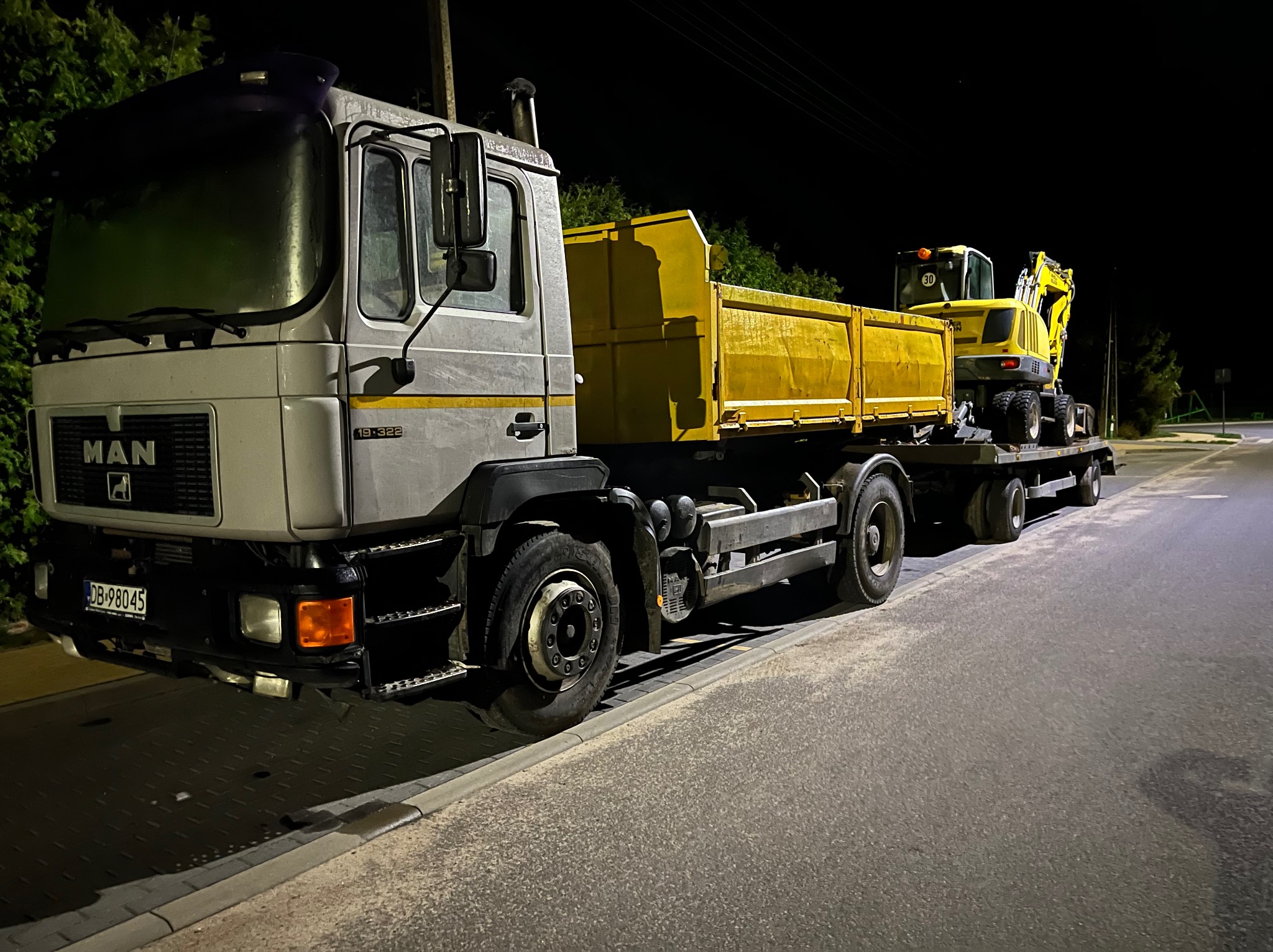 Ciężarówka MAN z żółtą naczepą przewożąca żółtą mini koparkę na przyczepie, nocne ujęcie.