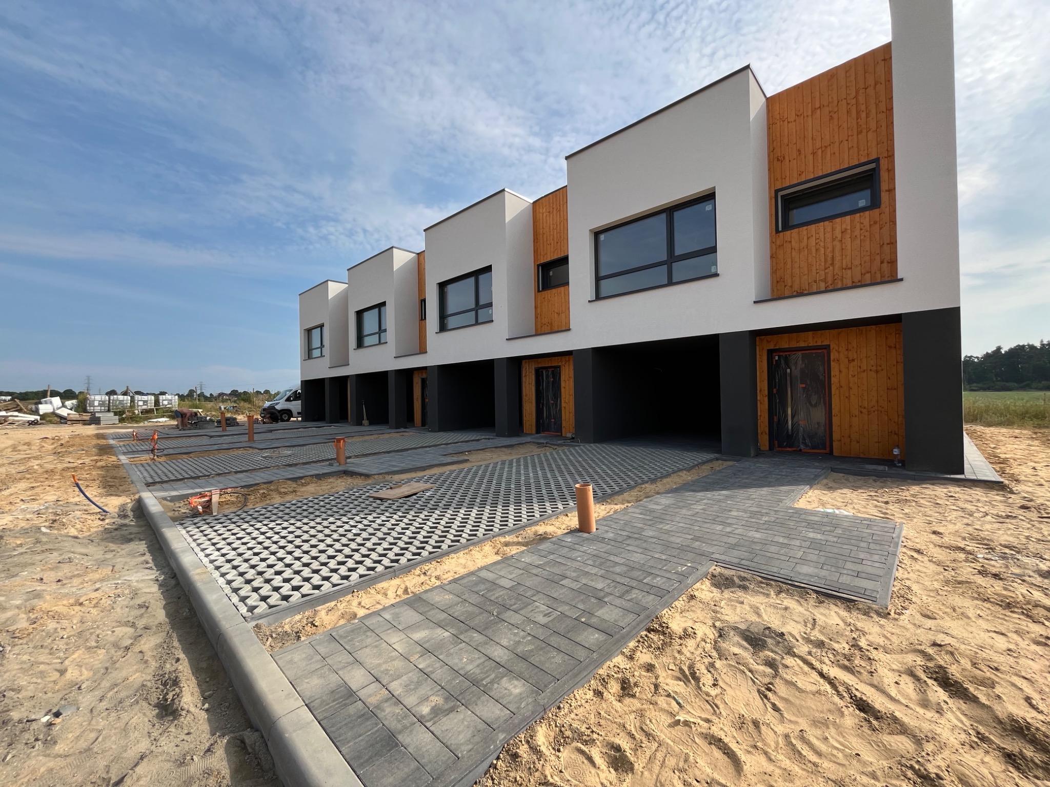 Układanie szarej kostki brukowej przed szeregiem nowoczesnych domów z elementami drewnianej elewacji, widoczne obrzeża i częściowo ułożona nawierzchnia ażurowa.