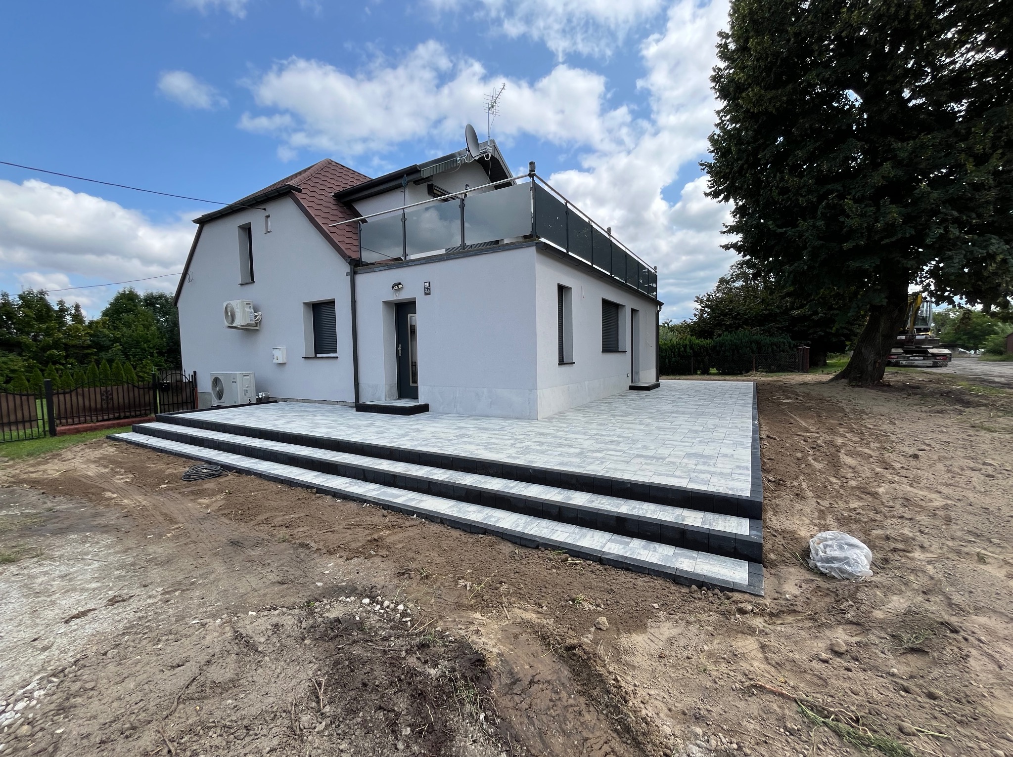 Nowoczesny dom z jasnoszarą elewacją i tarasem z szarej kostki brukowej, widoczne schody z czarnymi obrzeżami, ziemia w stanie przygotowania, w tle budynek i drzewa.