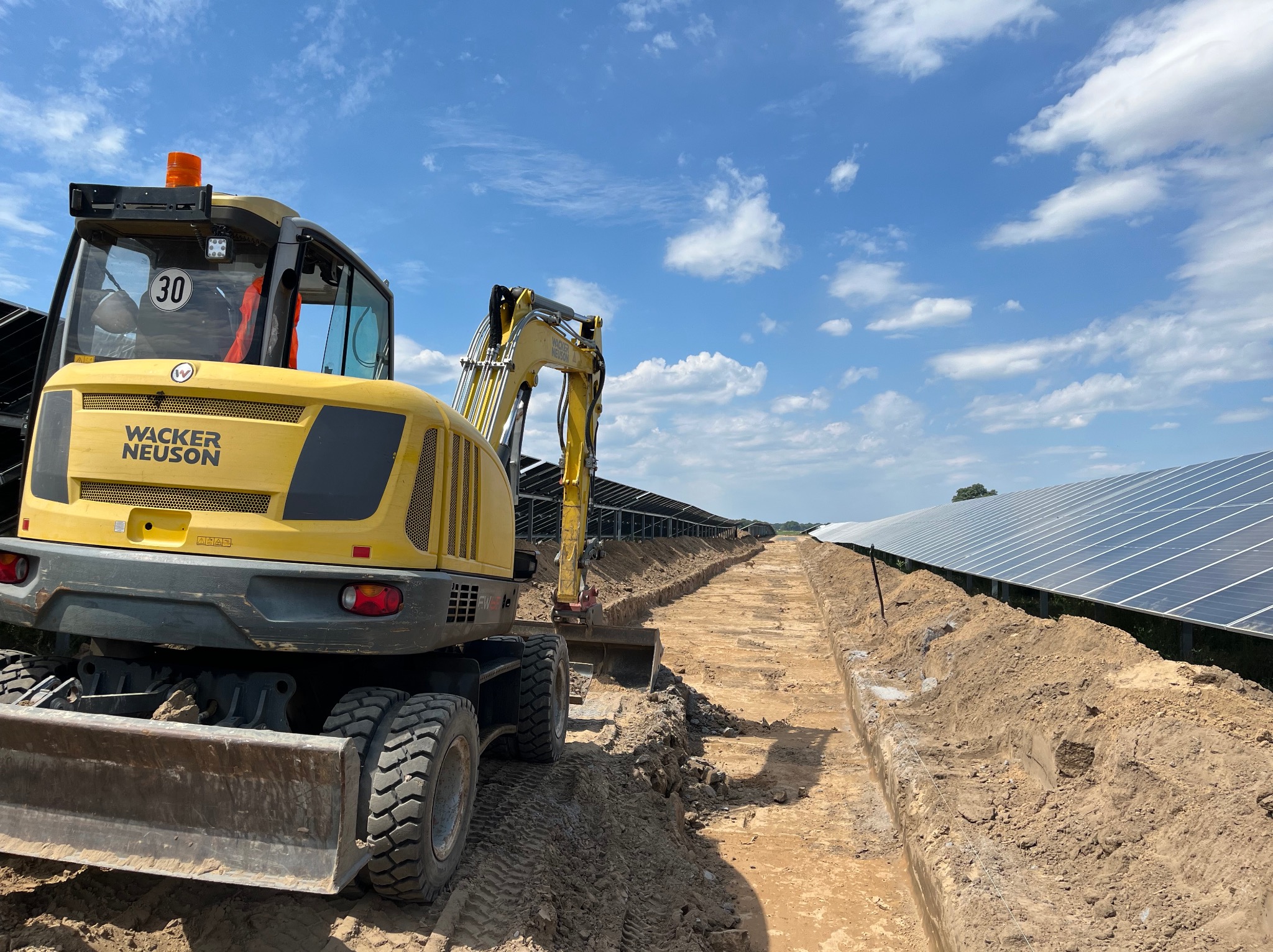 Żółta mini koparka Wacker Neuson podczas prac ziemnych, wykopany wąski rów pod panele słoneczne, błękitne niebo z chmurami w tle.