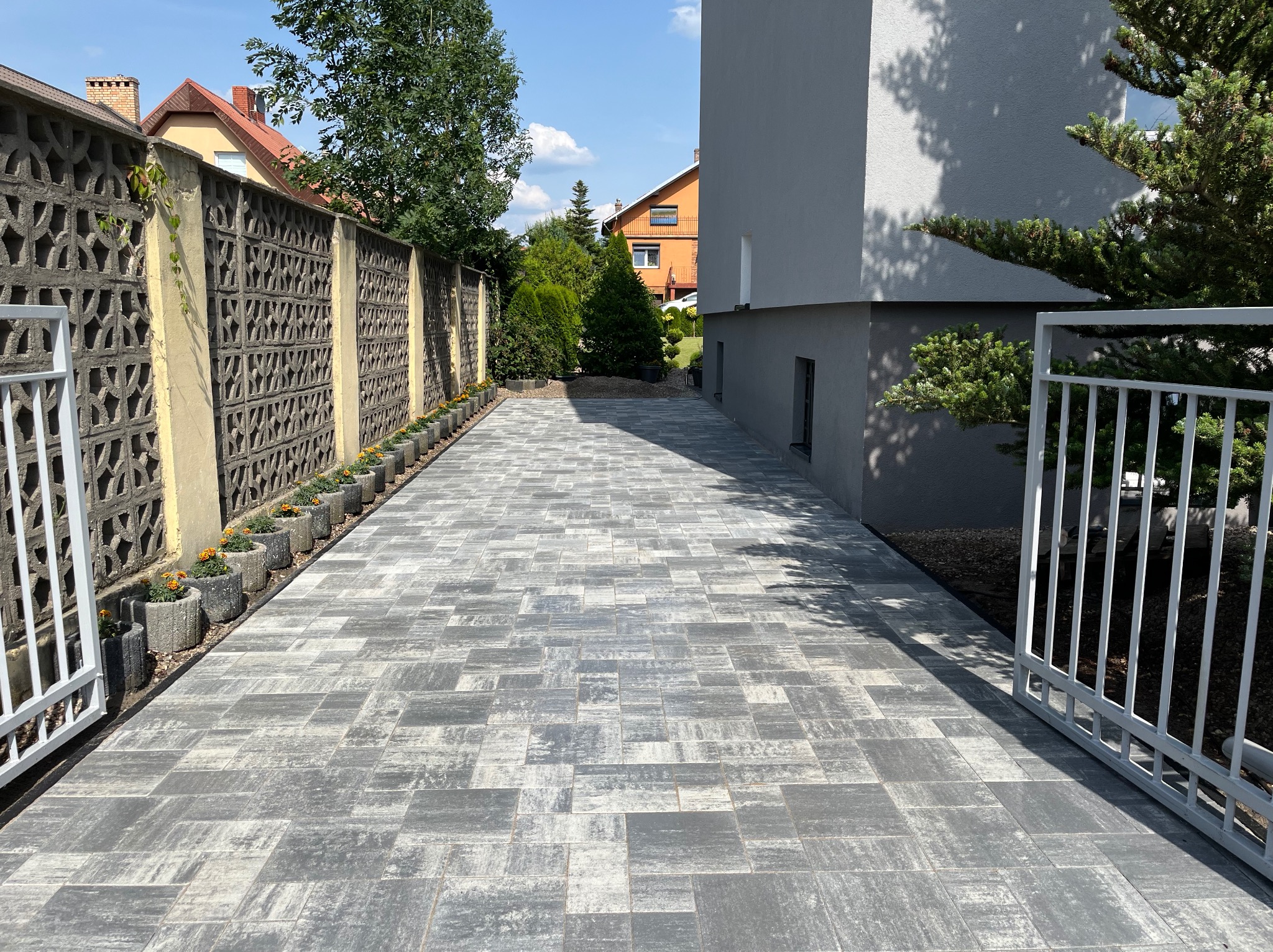 Nowo ułożona, szara kostka brukowa na podjeździe, z rzędem betonowych donic z pomarańczowymi kwiatami wzdłuż ażurowego ogrodzenia.