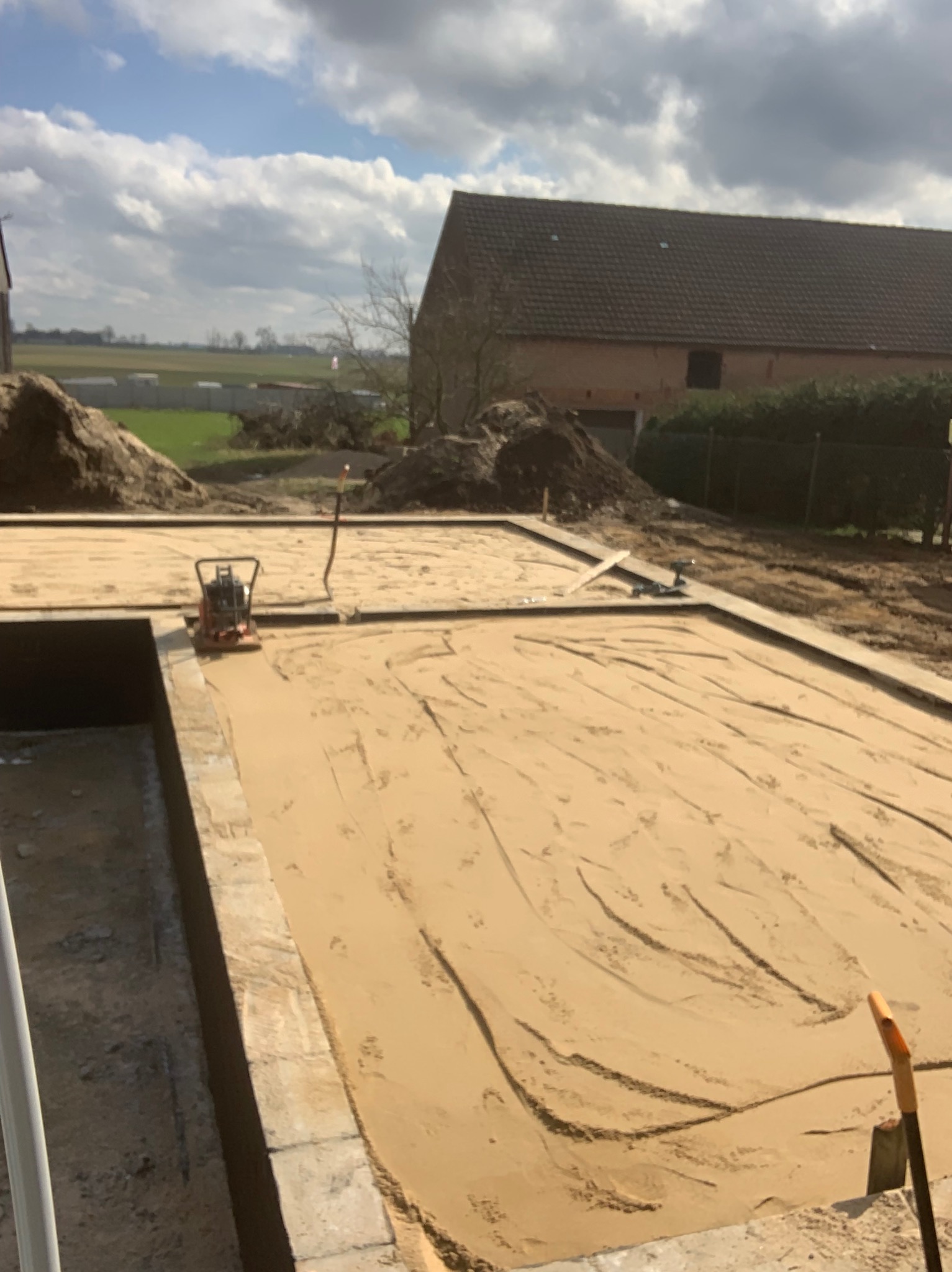 Przygotowany teren pod budowę z ubitym piaskiem, widoczny ubijak wibracyjny, obrzeża i pryzmy ziemi w tle, w oddali budynek mieszkalny i pola.