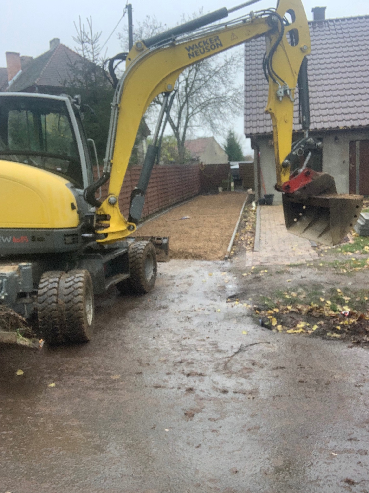 Żółta koparka Wacker Neuson na błotnistym terenie z przygotowanym podłożem pod kostkę brukową w tle, jesienne liście na ziemi.