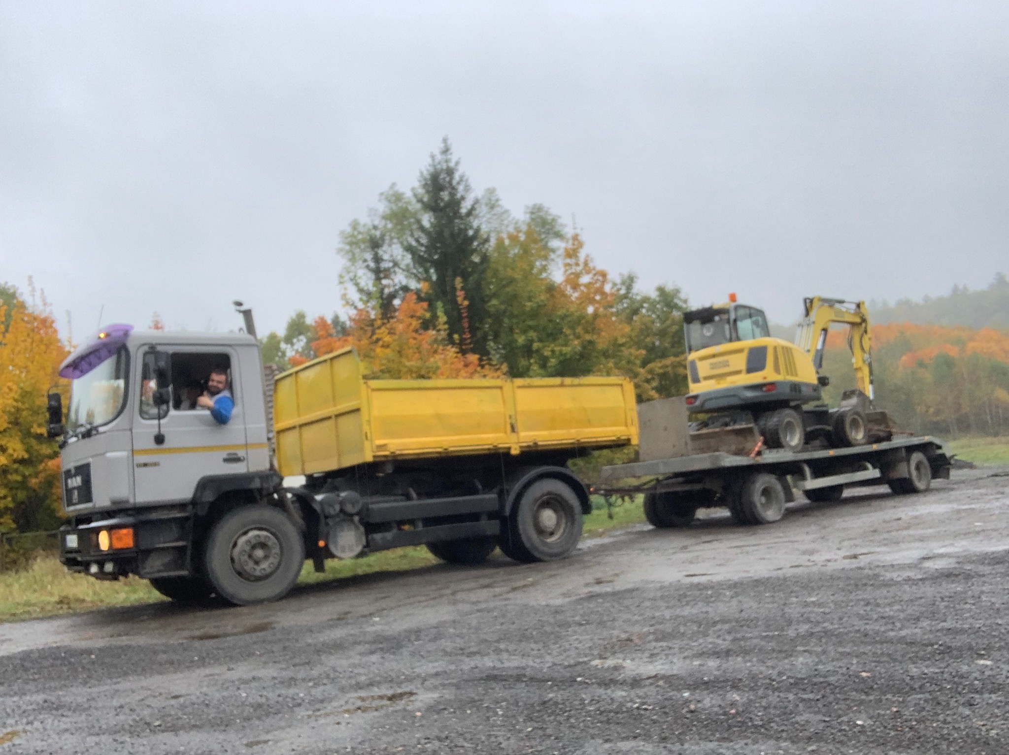 Ciężarówka MAN z żółtą skrzynią ładunkową ciągnąca lawetę z żółtą koparką kołową na tle jesiennego krajobrazu.