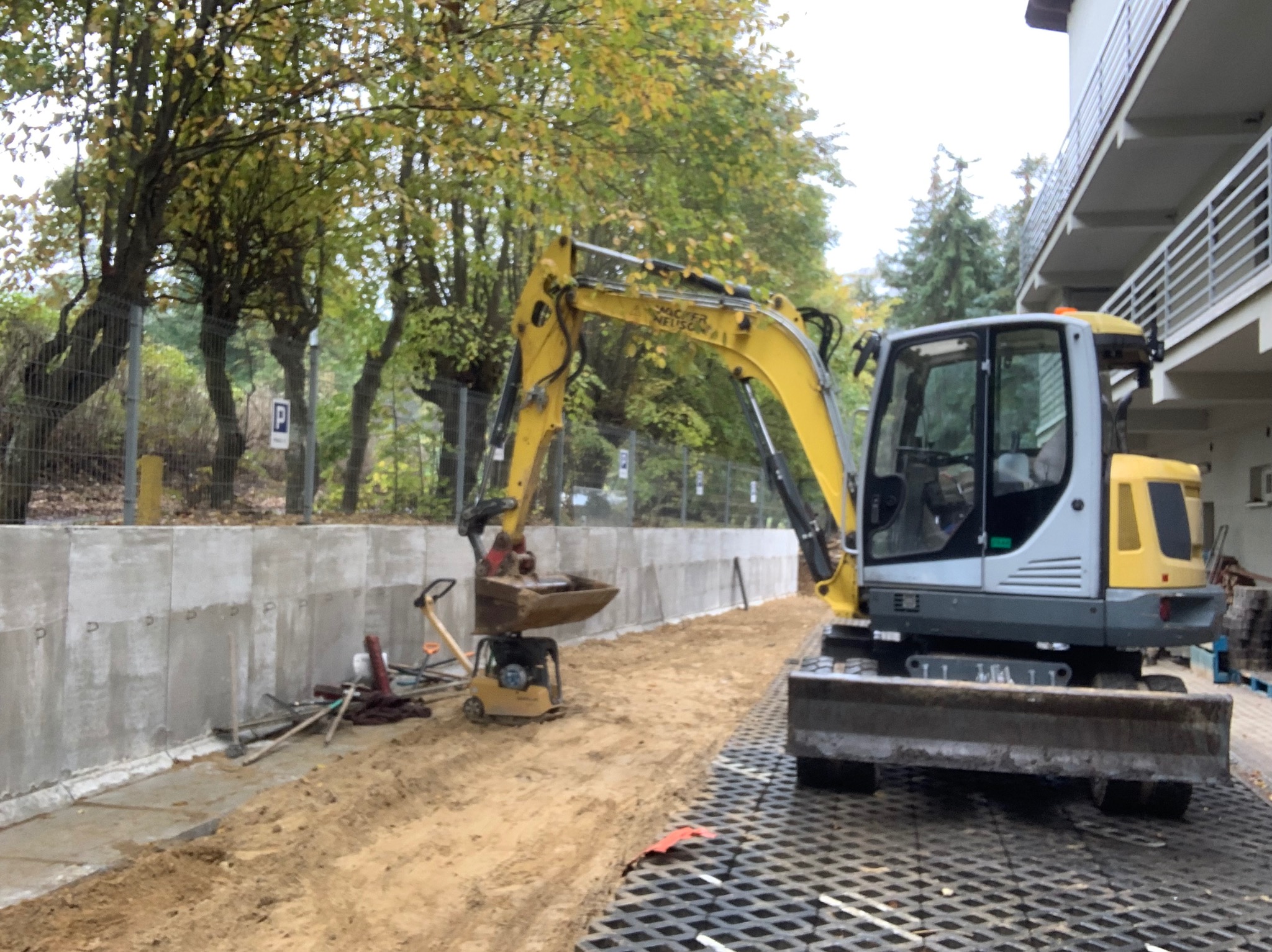 Żółto-szara mini koparka na placu budowy, obok betonowego muru oporowego i ubijarki, w tle drzewa i budynek z balkonami.