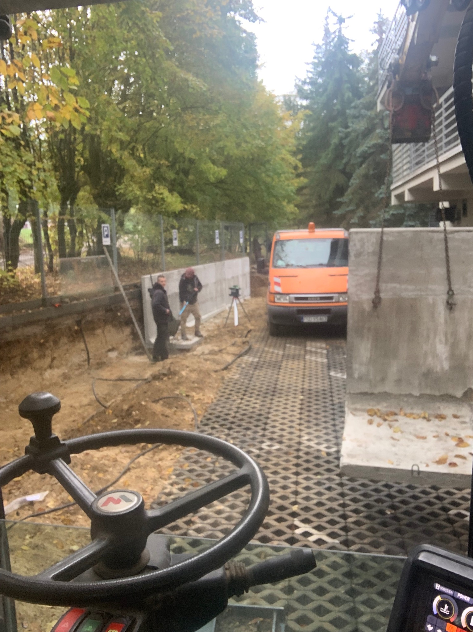 Widok z kabiny maszyny na plac budowy z wykopem, pracownikami i pomarańczowym samochodem dostawczym na tle drzew i budynku, widoczne betonowe elementy konstrukcyjne.