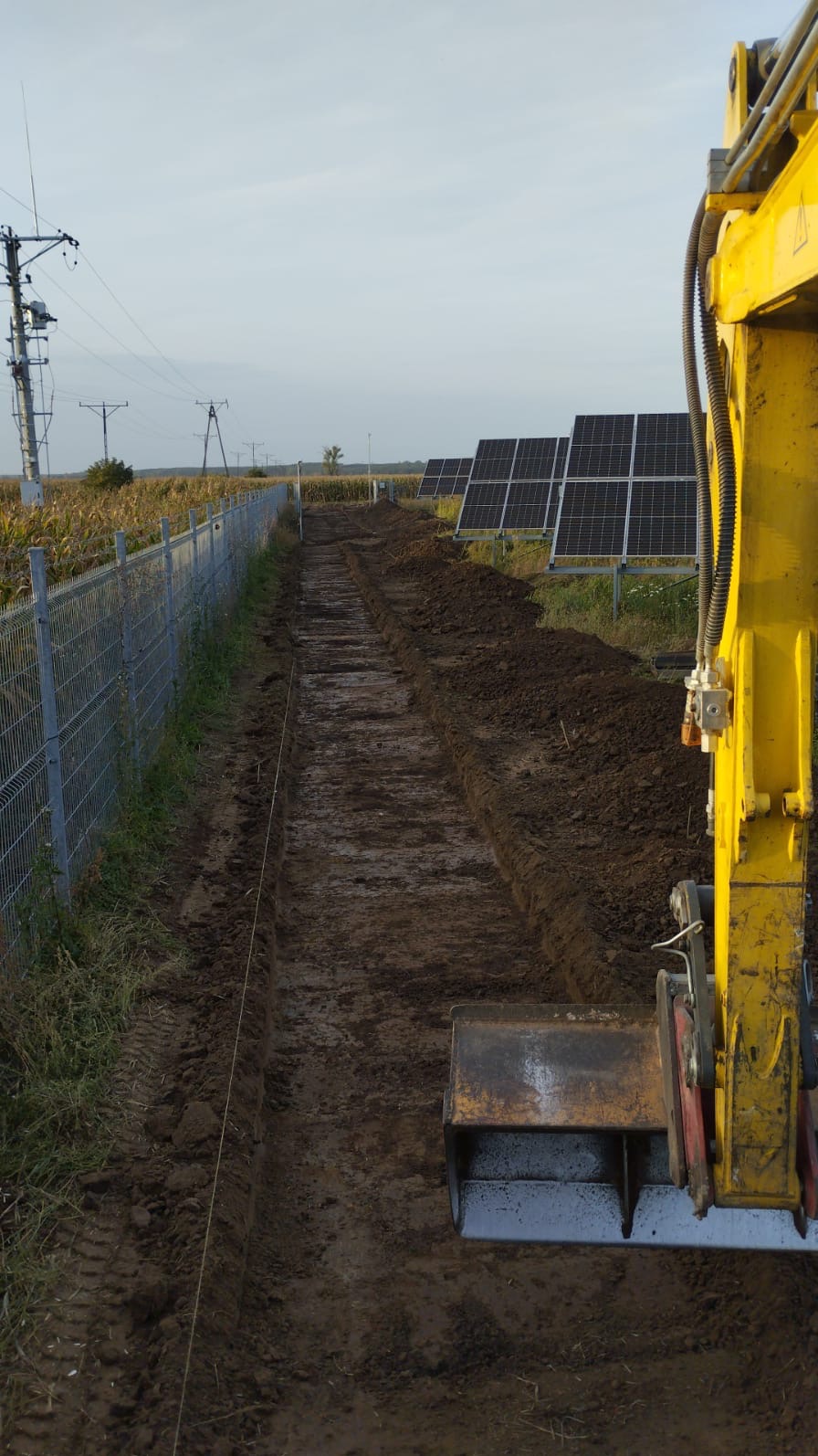 Żółta koparka wykonuje wąski wykop w ziemi wzdłuż ogrodzenia, w tle panele słoneczne i pole kukurydzy.