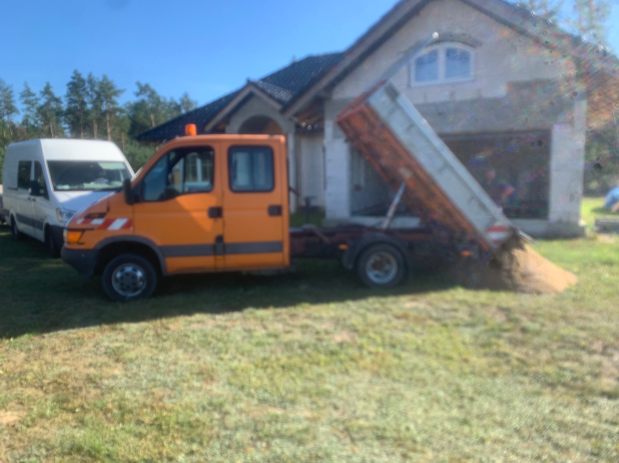 Pomarańczowa wywrotka z podniesioną skrzynią ładunkową, wysypująca ziemię na trawnik przed budynkiem w trakcie słonecznego dnia; obok zaparkowany biały bus.