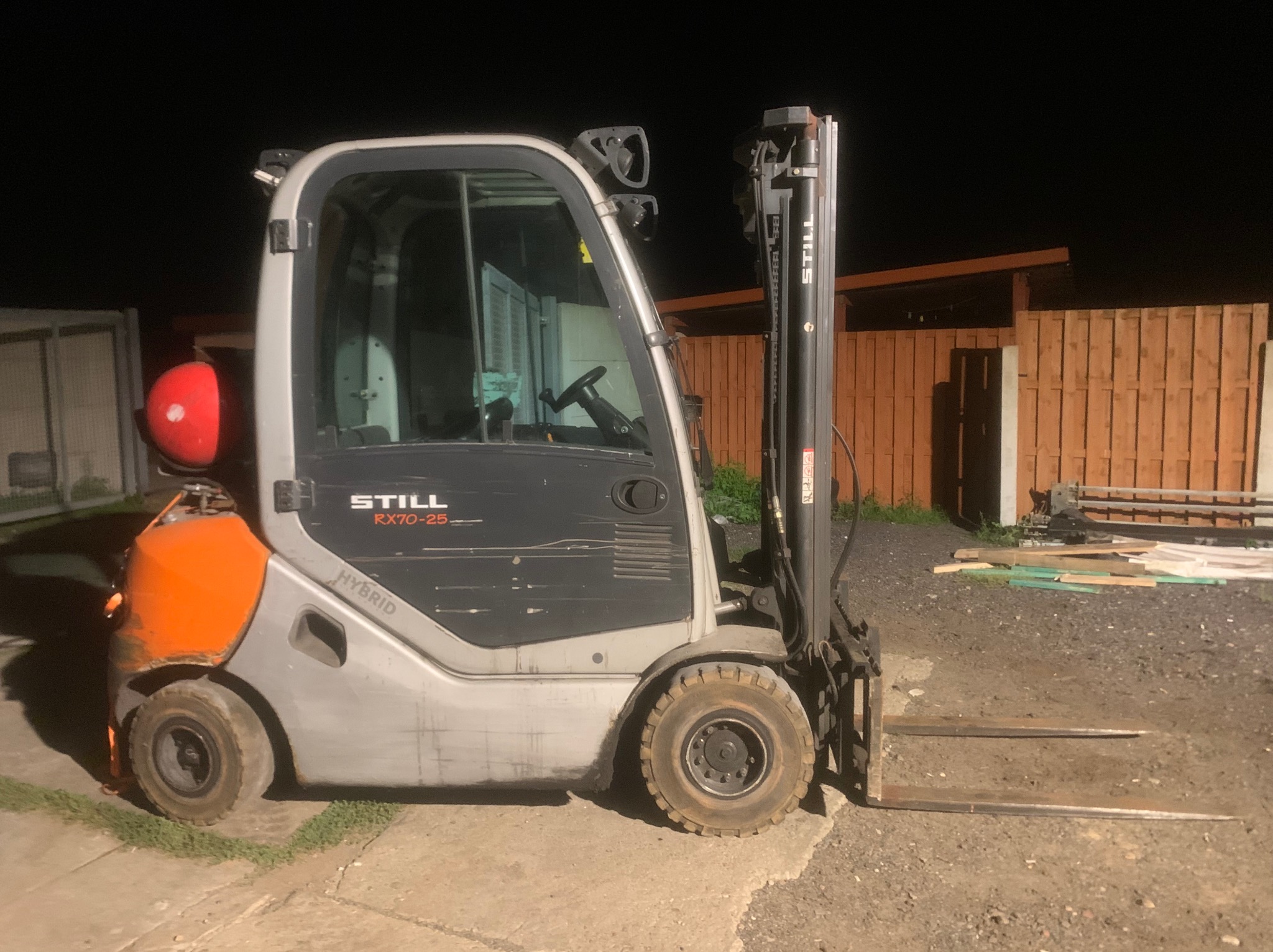 Hybrydowy wózek widłowy STILL RX70-25 z widocznymi śladami użytkowania, zaparkowany na utwardzonym terenie, z drewnianym ogrodzeniem w tle.