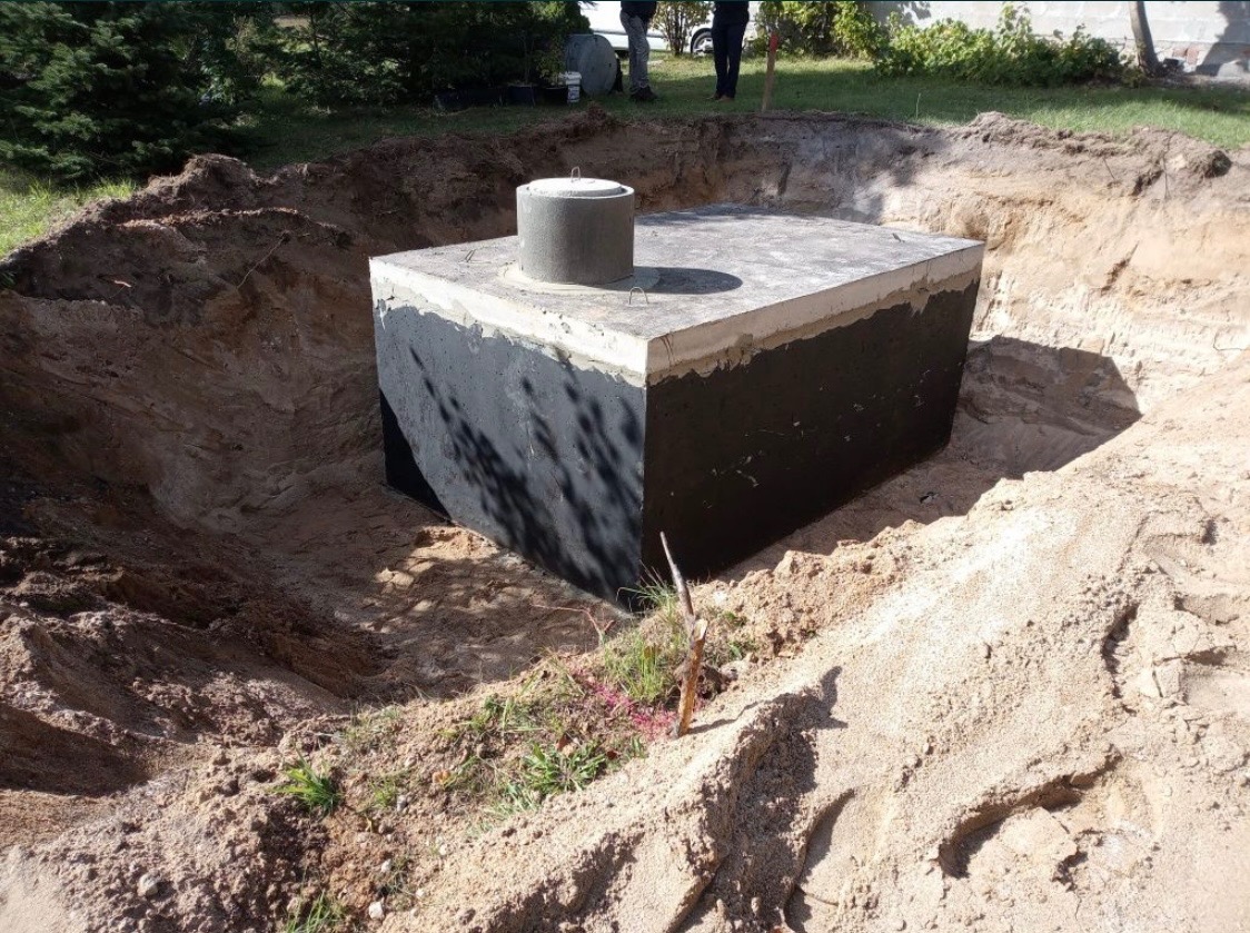 Betonowy zbiornik z pokrywą, częściowo zagłębiony w ziemi w Strzelcach Krajeńskich, otoczony wykopem z piasku i ziemi, widoczne ślady świeżych prac ziemnych.