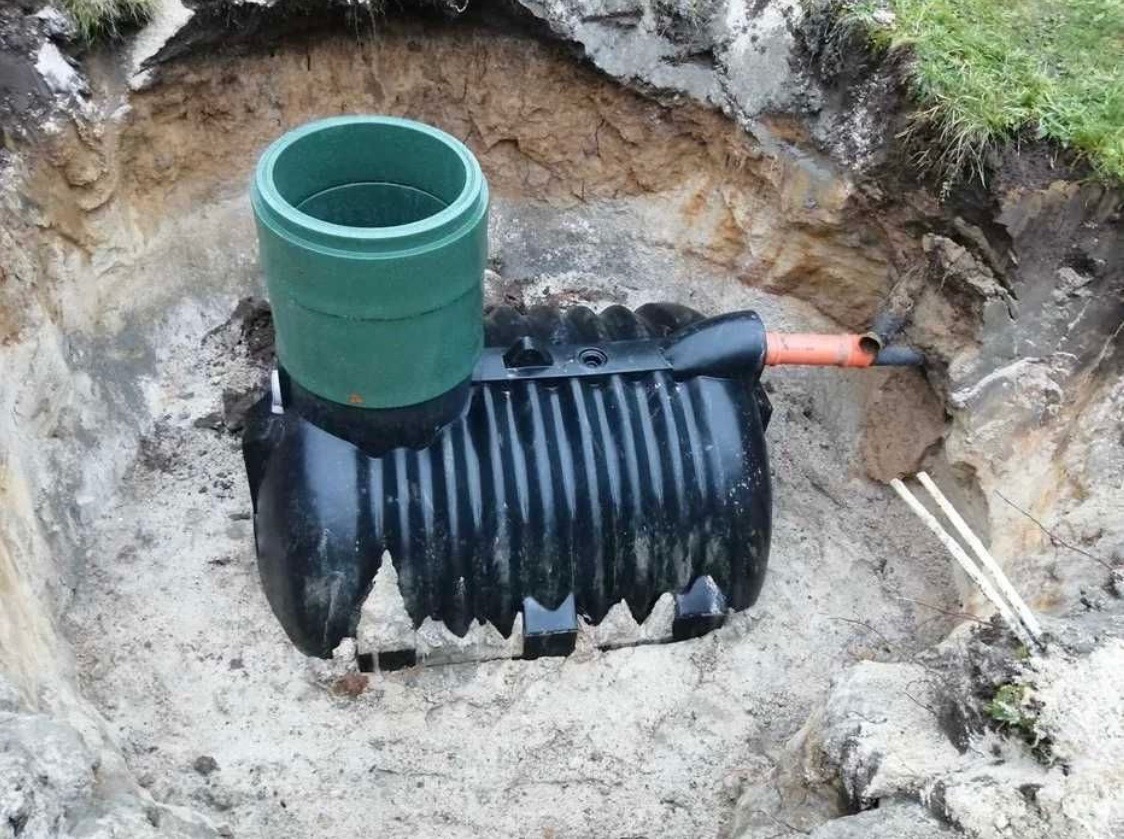 Osadnik gnilny z zielonym kominkiem wentylacyjnym i pomarańczową rurą odprowadzającą, umieszczony w wykopie z piaskową ziemią, widok z góry.