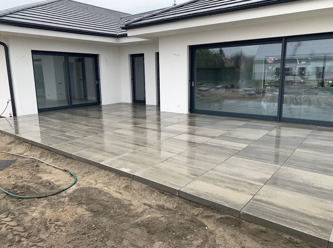 Świeżo ułożony, mokry taras z szarych, prostokątnych płyt imitujących kamień, przylegający do białego domu z dużymi przesuwnymi drzwiami tarasowymi i czarnym dachem, widoczny wąż ogrodowy na skraju.