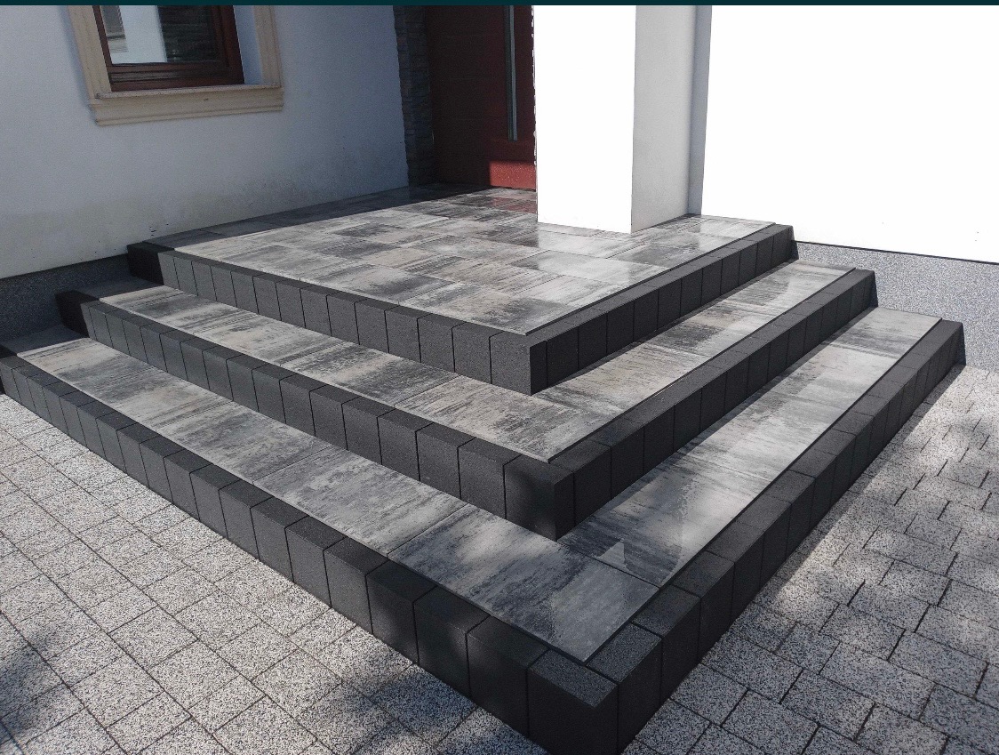 Schody wejściowe wyłożone szarą kostką brukową z ciemnymi obrzeżami, prowadzące do budynku z białymi ścianami i stolarką okienną w odcieniach beżu i brązu.