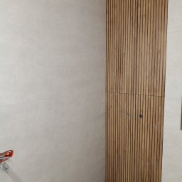 Mateusz Mateusz - Jasne płytki ścienne i panel z drewnianych listew z ukrytymi drzwiczkami rewizyjnymi w nowoczesnej łazience.