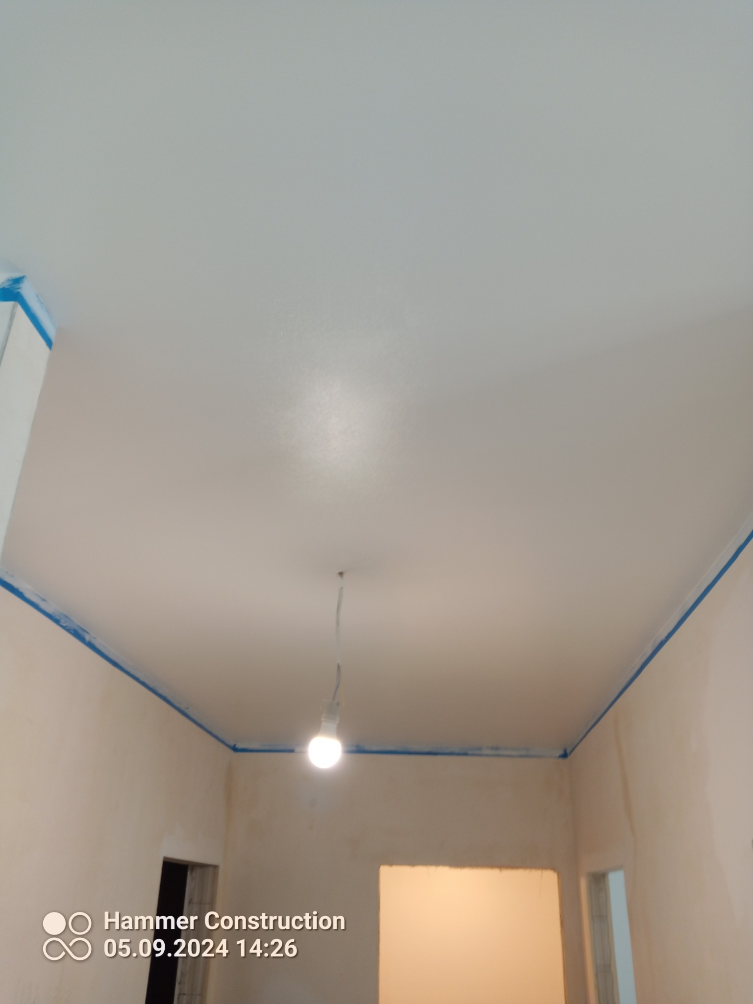Biały sufit po nałożeniu gładzi, zabezpieczony niebieską taśmą malarską przy ścianach, z widoczną żarówką na kablu.