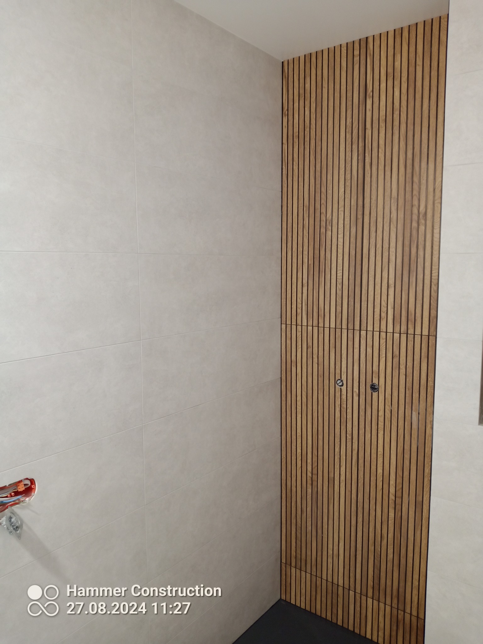 Jasne płytki ścienne i panel z drewnianych listew z ukrytymi drzwiczkami rewizyjnymi w nowoczesnej łazience.