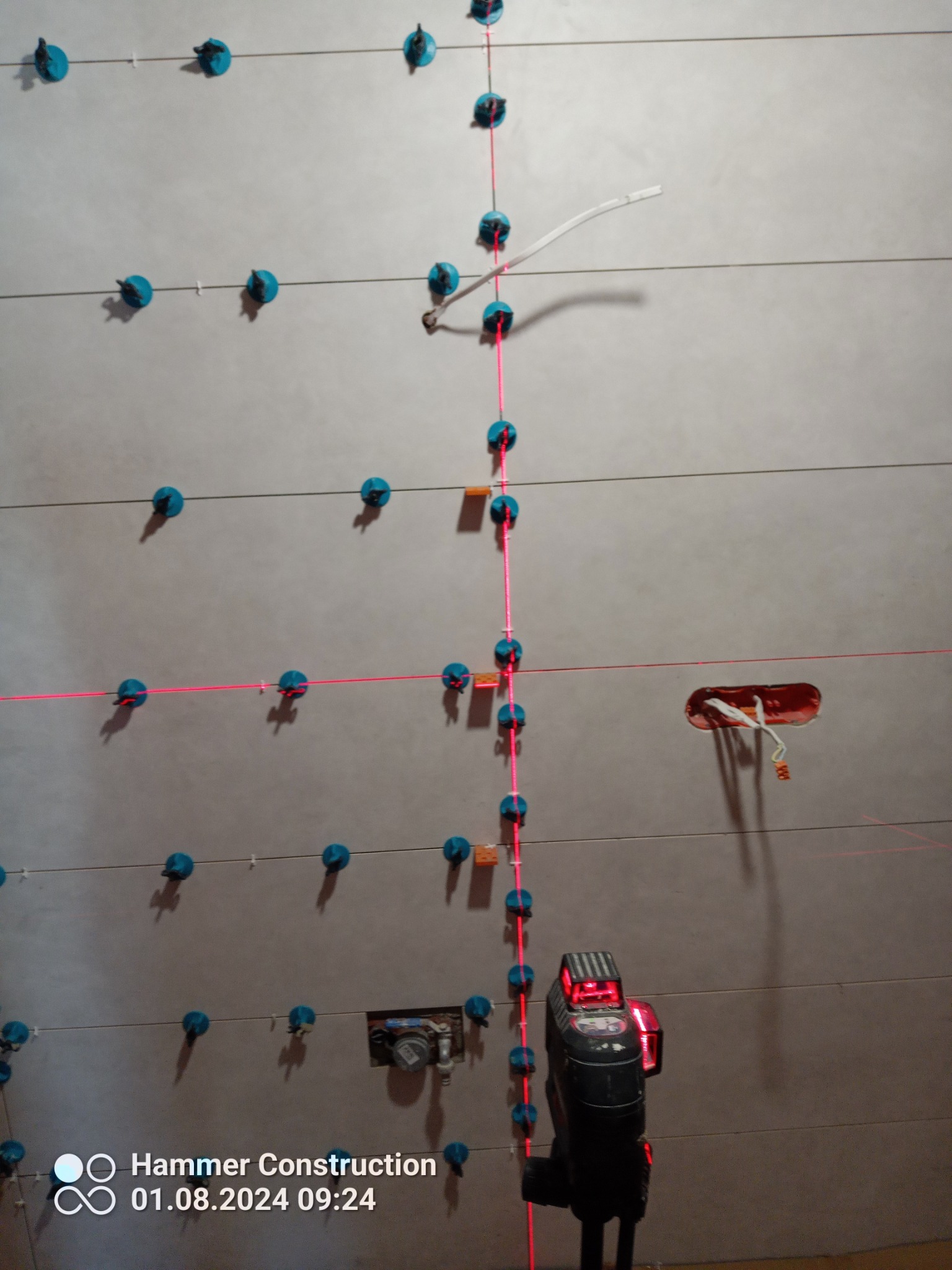 Układanie płytek na ścianie z użyciem poziomicy laserowej i systemu poziomowania płytek z niebieskimi klipsami, widoczne gniazdko elektryczne z wystającymi kablami.