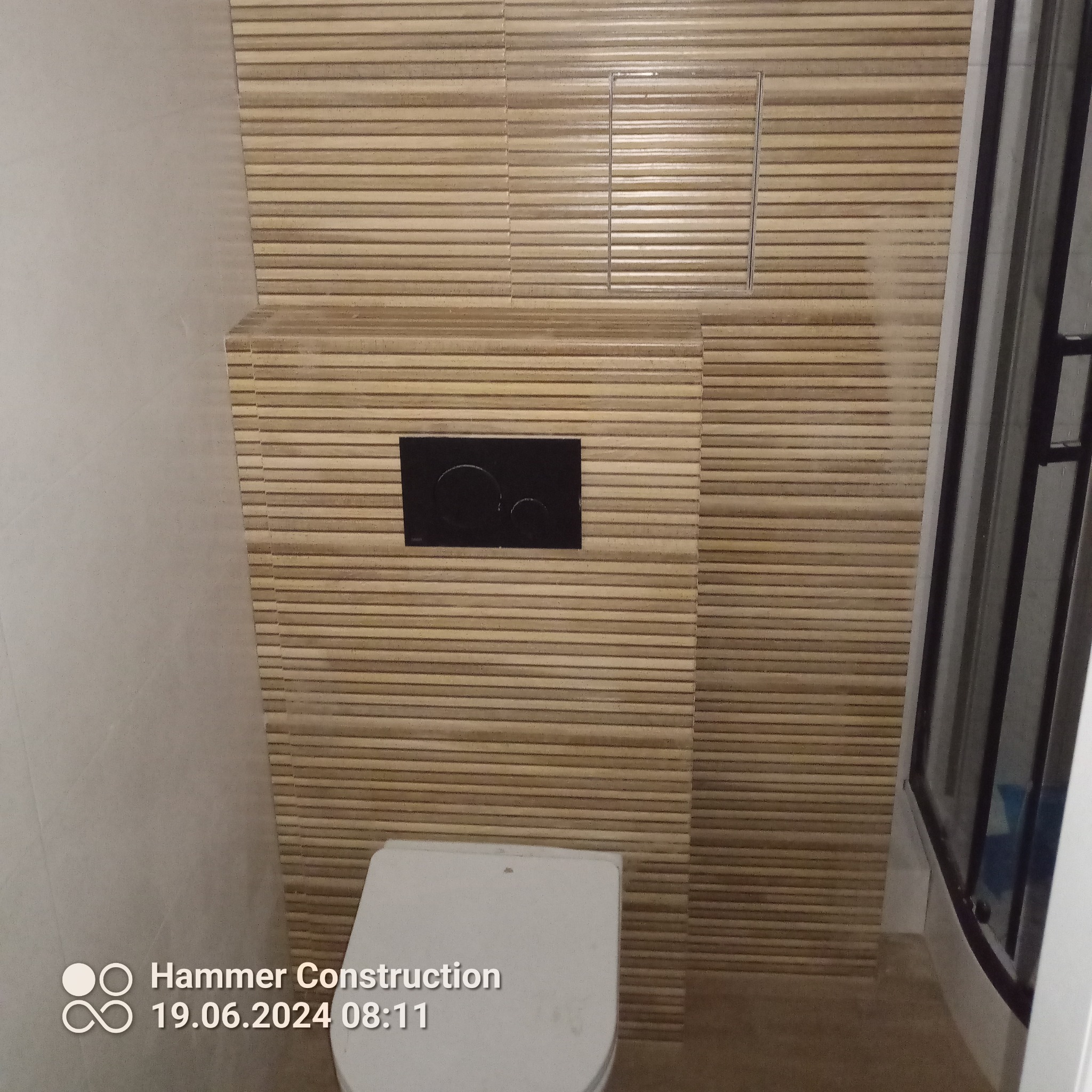 Wykończona toaleta z poziomymi płytkami imitującymi drewno, czarny przycisk spłuczki i częściowo widoczna kabina prysznicowa po prawej.