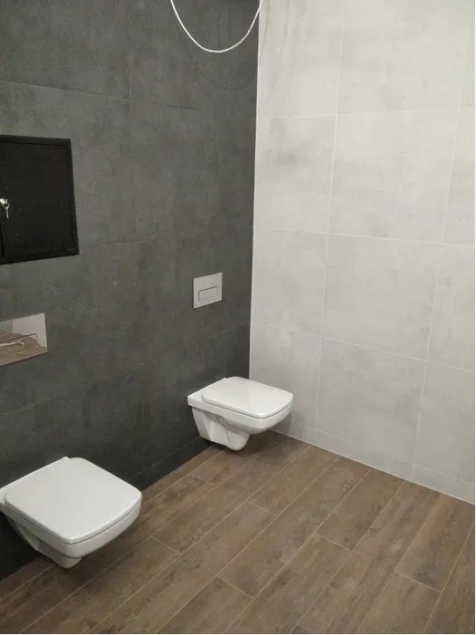 Nowoczesna łazienka z dwoma podwieszanymi toaletami, ciemnoszara ściana z czarną szafką rewizyjną, jasnoszara ściana z płytkami oraz podłoga imitująca drewno.