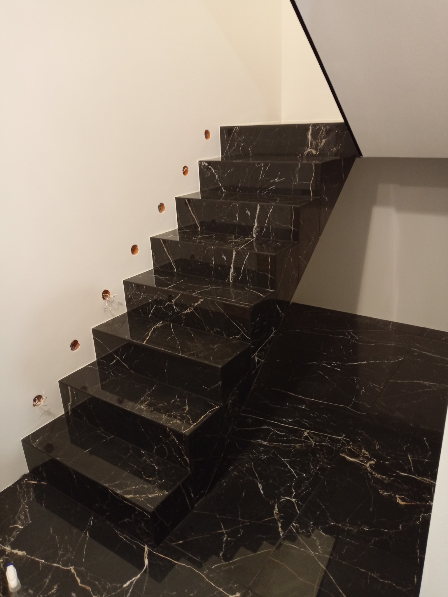 Schody wyłożone czarnym marmurem z białymi żyłkami, widoczne otwory na oświetlenie w ścianie, minimalistyczny design.