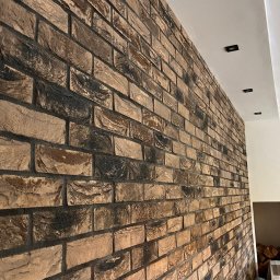 Adam Kostecki Remonty Szczecin - Ściana wykończona dekoracyjną cegłą w odcieniach brązu i czerni, z widocznymi fugami i minimalistycznym oświetleniem sufitowym.