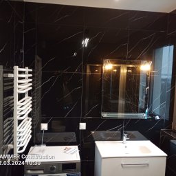 Adam Kostecki Remonty Szczecin - Nowoczesna łazienka z czarnymi płytkami imitującymi marmur, białą umywalką i grzejnikiem drabinkowym. Widoczna pralka z ulotką reklamową na górze. Oświetlenie sufitowe odbija się w lustrze.