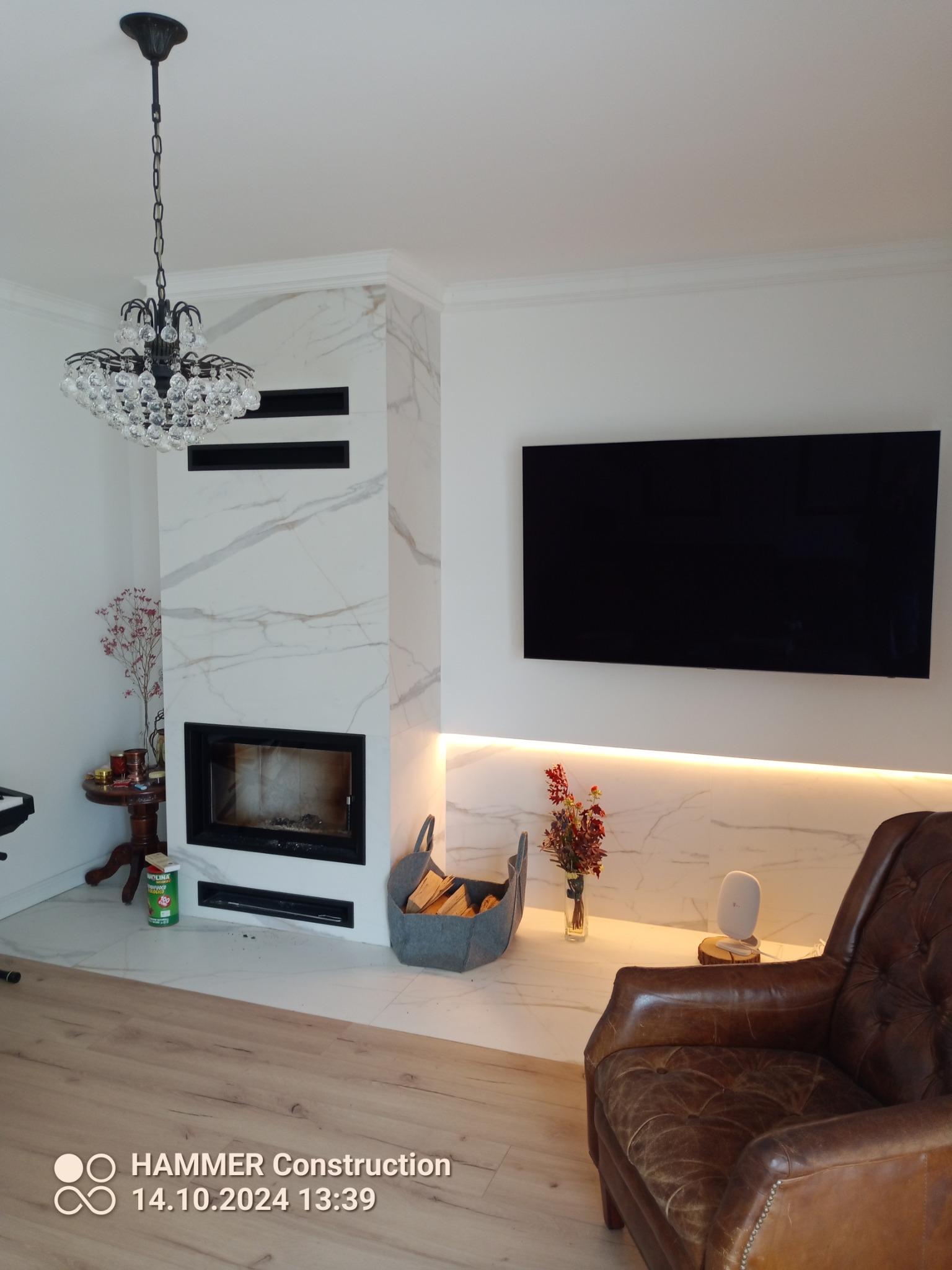 Salon z marmurowym kominkiem, wiszącym telewizorem, kryształowym żyrandolem i skórzanym fotelem w stylu vintage.