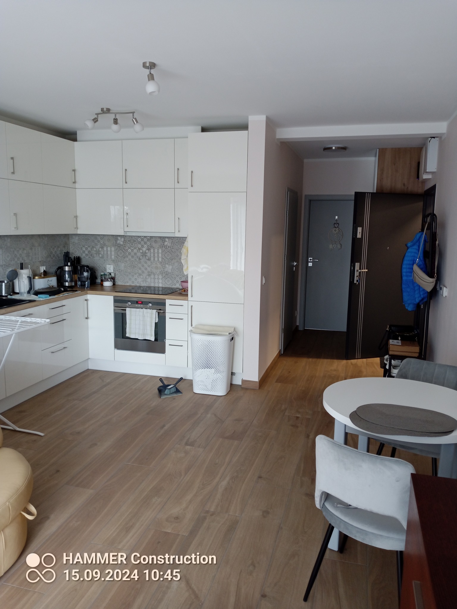 Jasna kuchnia z białymi meblami i drewnopodobną podłogą, widok na stół z krzesłami oraz fragment przedpokoju z drzwiami wejściowymi.