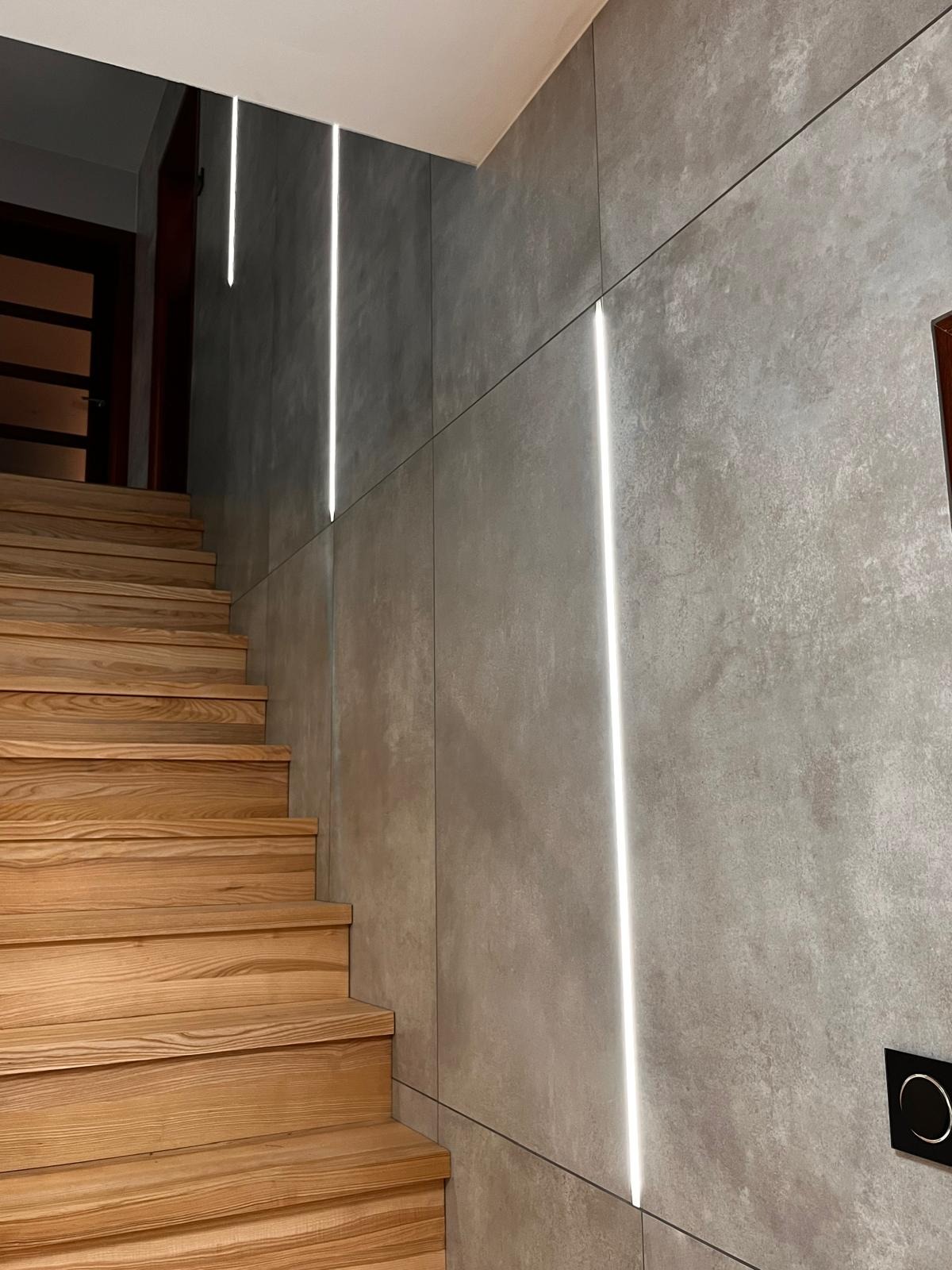 Nowoczesne schody drewniane z podświetlanymi pionowymi panelami ściennymi w odcieniach szarości, minimalistyczny design.