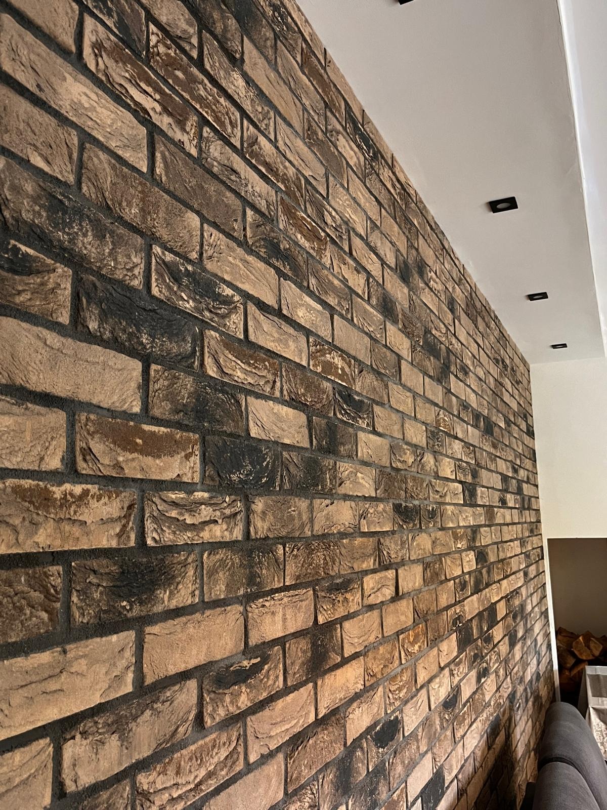 Ściana wykończona dekoracyjną cegłą w odcieniach brązu i czerni, z widocznymi fugami i minimalistycznym oświetleniem sufitowym.