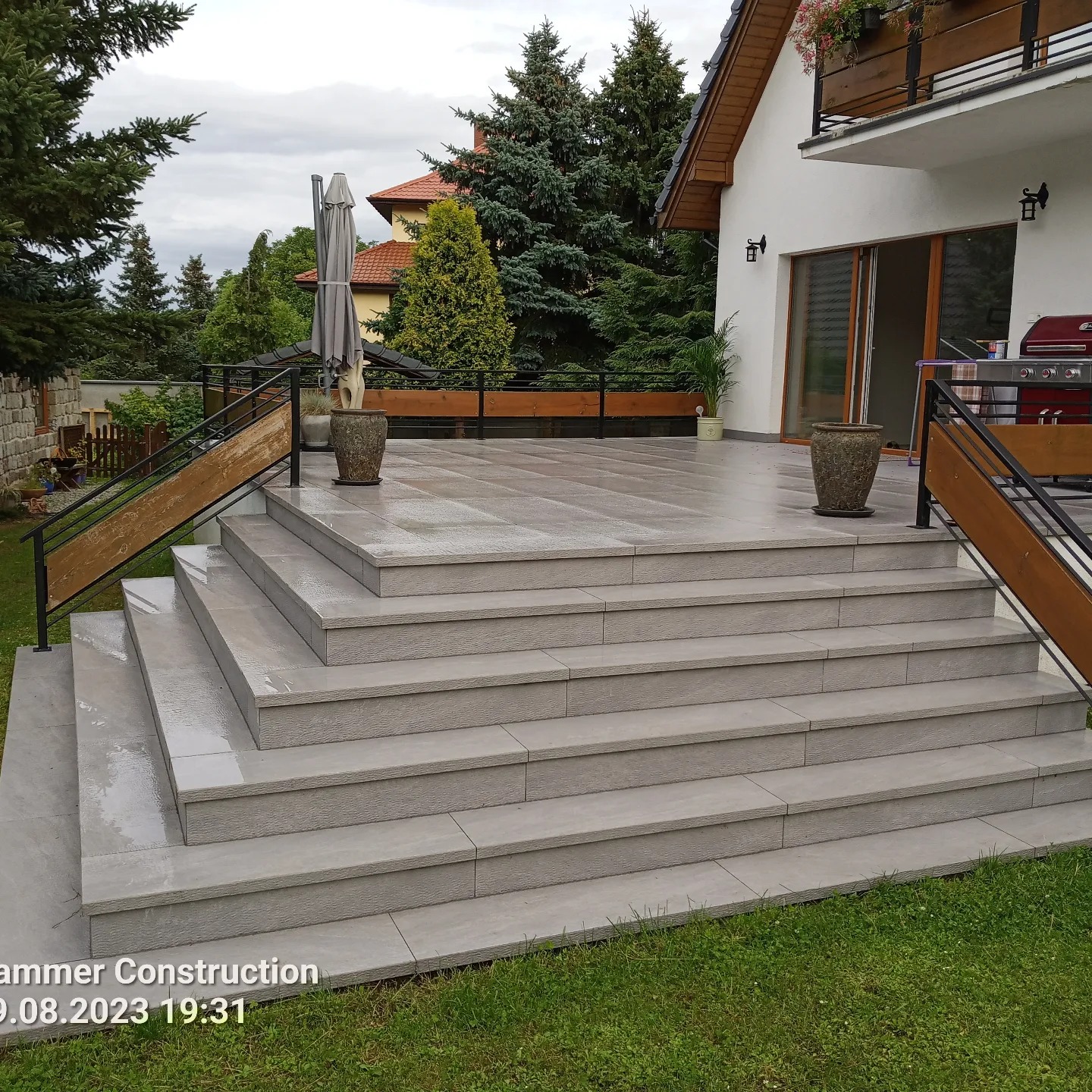 Schody z szarych płyt prowadzące na taras z drewnianą balustradą przy białym domu z balkonem i zielonym ogrodem w tle, po deszczu.