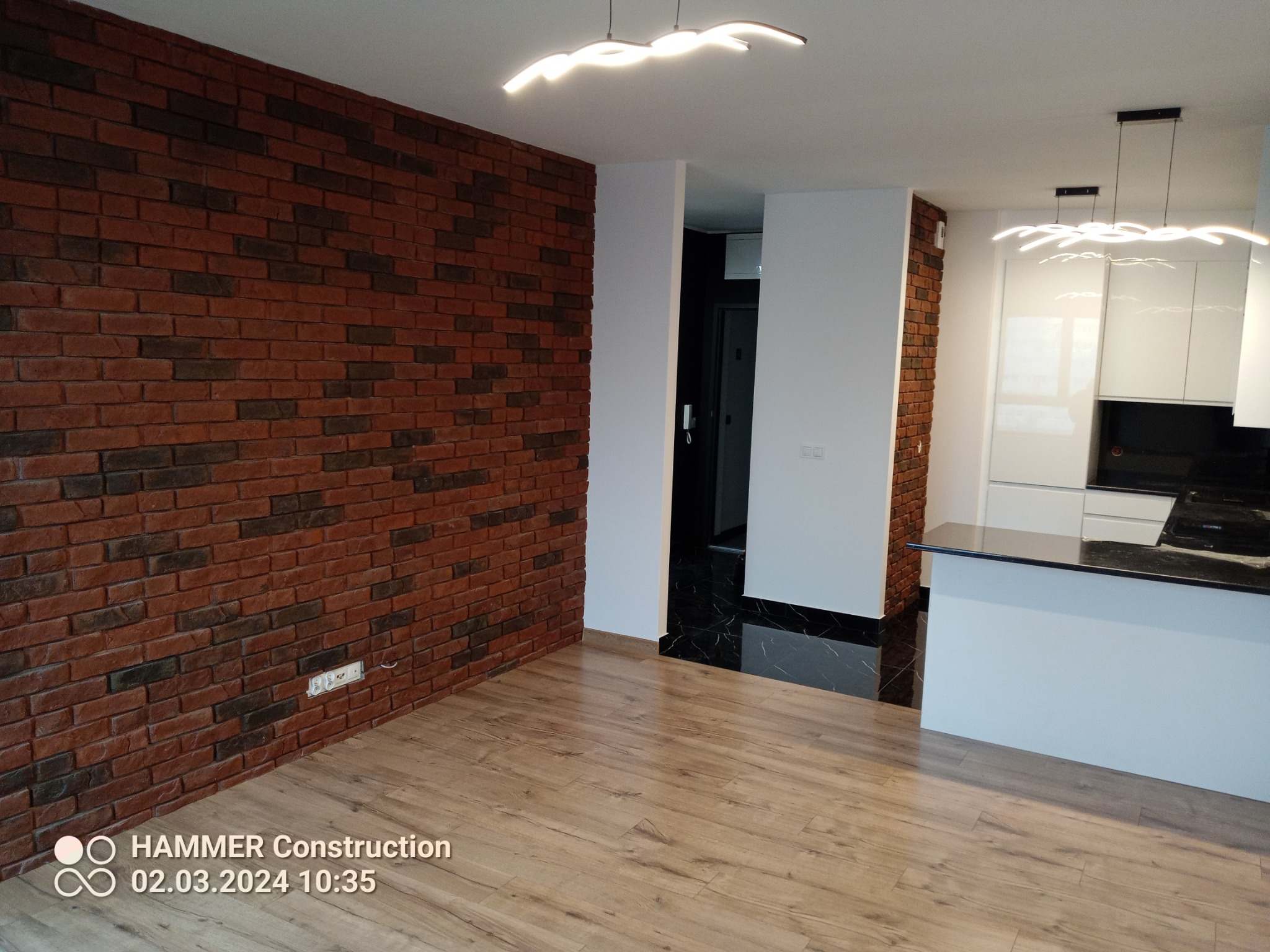 Salon z ceglaną ścianą, nowoczesnym oświetleniem i drewnianą podłogą, widok na kuchnię z białymi szafkami i czarnym blatem.