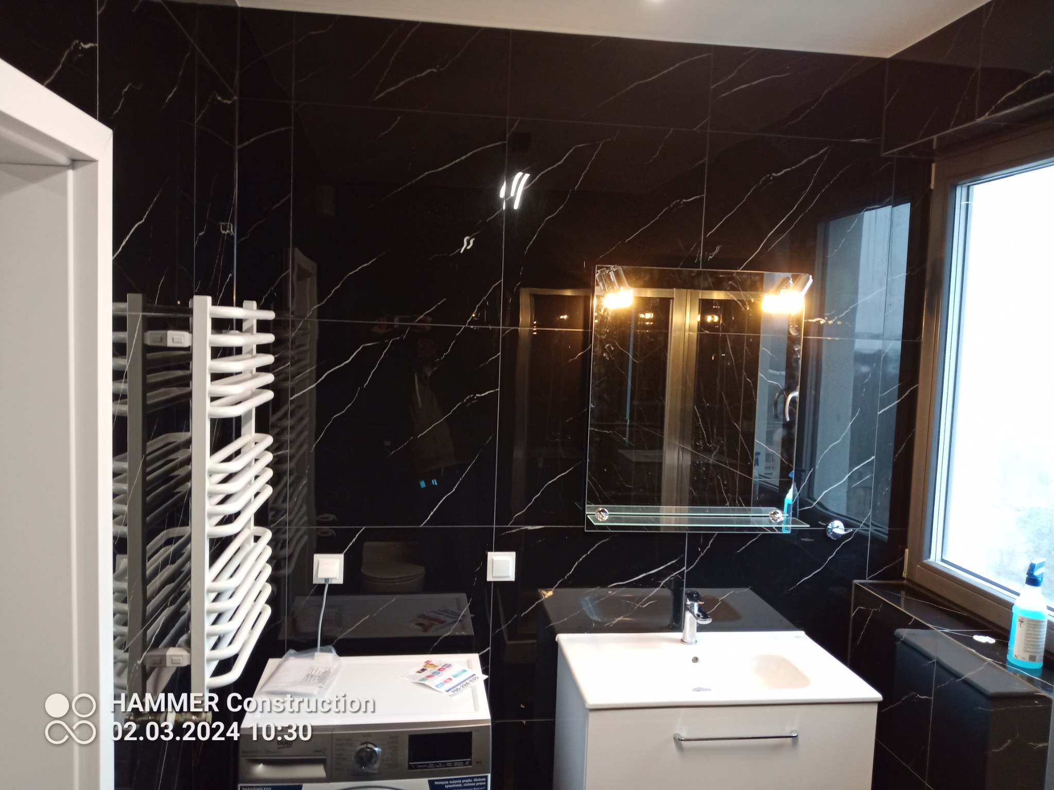 Nowoczesna łazienka z czarnymi płytkami imitującymi marmur, białą umywalką i grzejnikiem drabinkowym. Widoczna pralka z ulotką reklamową na górze. Oświetlenie sufitowe odbija się w lustrze.