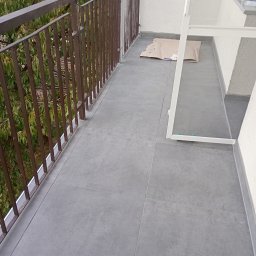 VT_PRO_MASTER - Szare płytki gresowe na balkonie z brązową balustradą, zabezpieczone kartonem przed malowaniem, widoczny fragment białej ramy okiennej.
