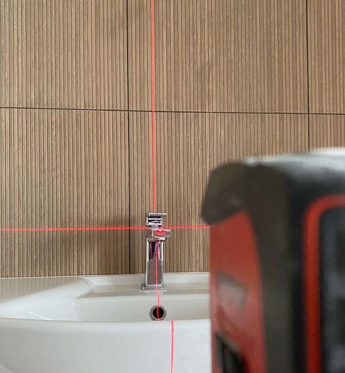 Montaż baterii umywalkowej z użyciem poziomicy laserowej; tło stanowią panele ścienne o fakturze drewna.