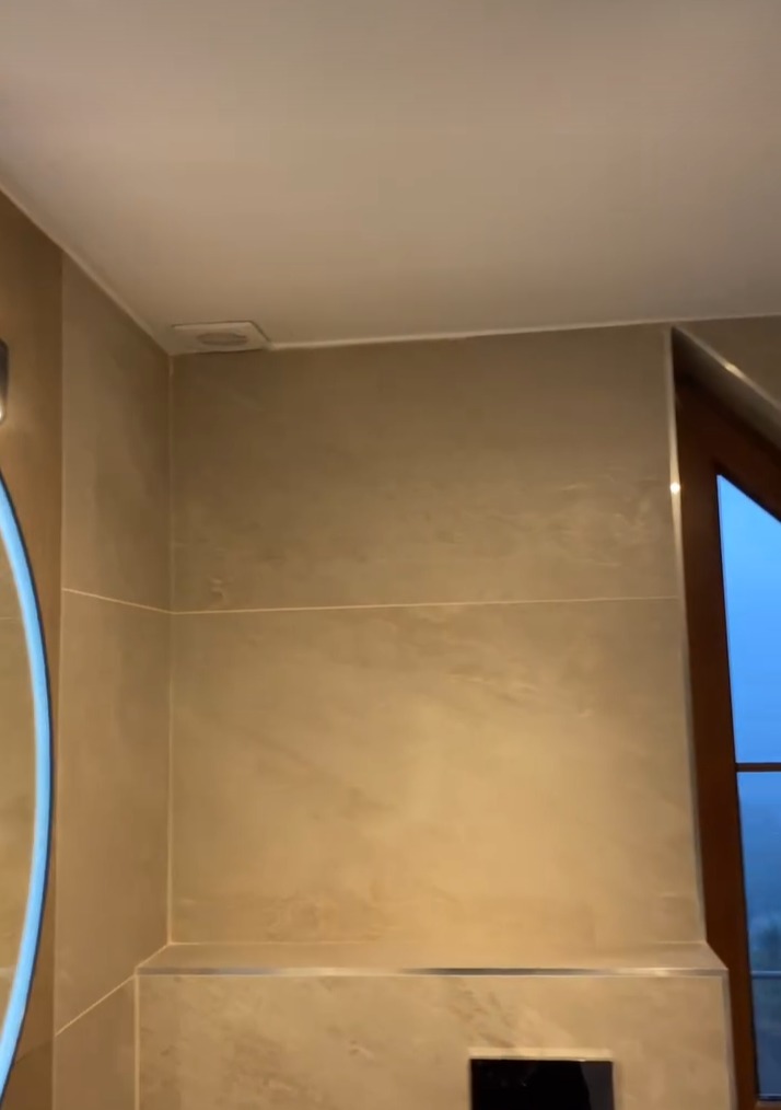 Fragment łazienki wykończonej w beżowych płytkach imitujących kamień, widoczny fragment podświetlanego lustra, okno z drewnianą ramą i kratka wentylacyjna pod sufitem.