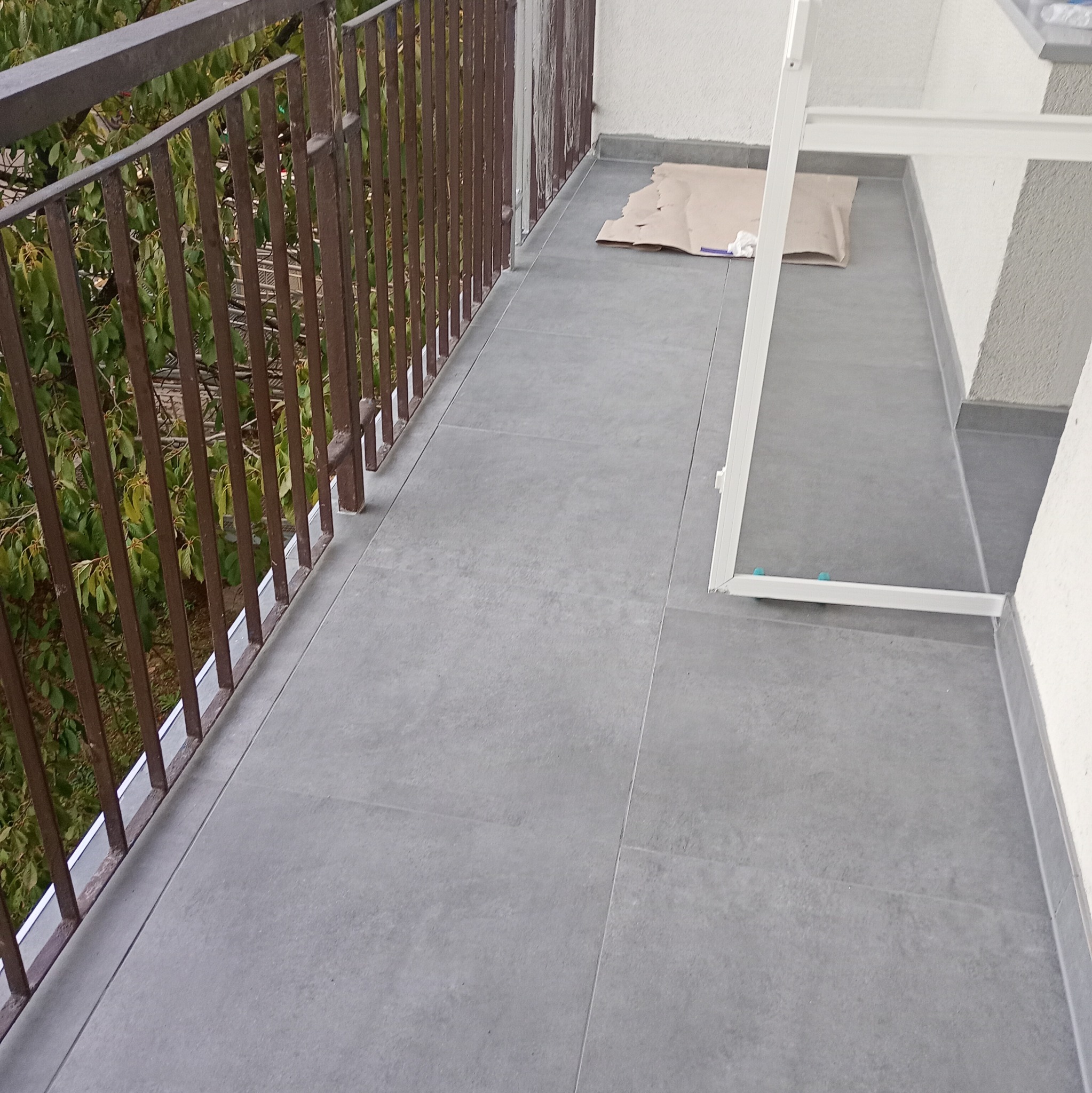 Szare płytki gresowe na balkonie z brązową balustradą, zabezpieczone kartonem przed malowaniem, widoczny fragment białej ramy okiennej.
